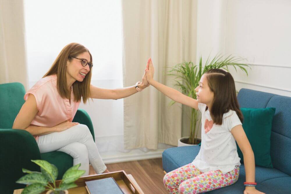 Erwachsene Frau und Mädchen geben sich High-Five in gemütlichem Raum mit Pflanzen, lächelnd und entspannt.