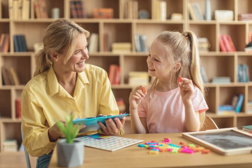 Frau und Mädchen sitzen am Tisch mit Spielzeug, Bücherregal im Hintergrund, beide lächeln sich an.