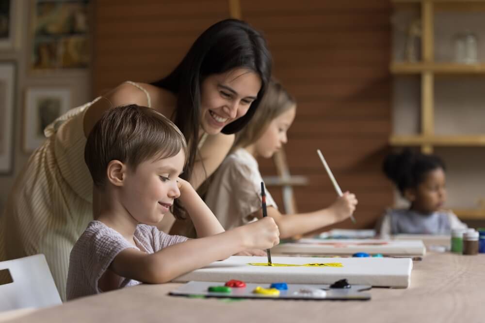 Lächelnde Erwachsene unterstützt Kinder beim Malen an einem Tisch in kreativem Umfeld.