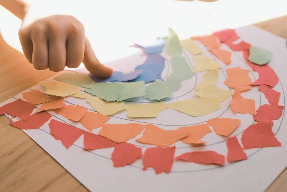Kind gestaltet bunte Collage aus gerissenen Papierstücken auf einem Tisch.