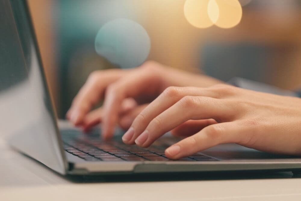 Nahaufnahme von Händen, die auf einem Laptop tippen, verschwommener Hintergrund mit warmen Lichtern.