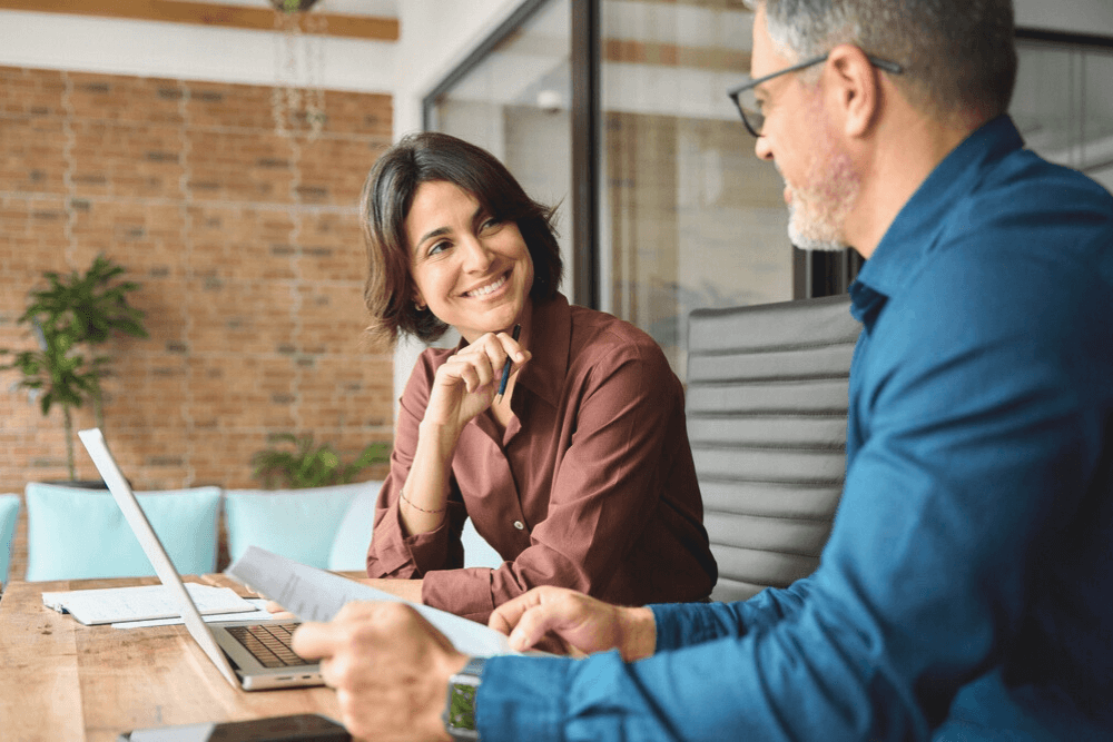 Zwei Personen diskutieren lächelnd in einem modernen Büro mit Laptop und Dokumenten.
