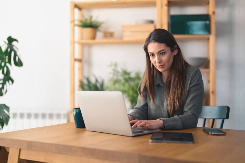 Frau am Laptop in modernem Büro mit Pflanzen und Holzmöbeln, konzentriert am Arbeiten.